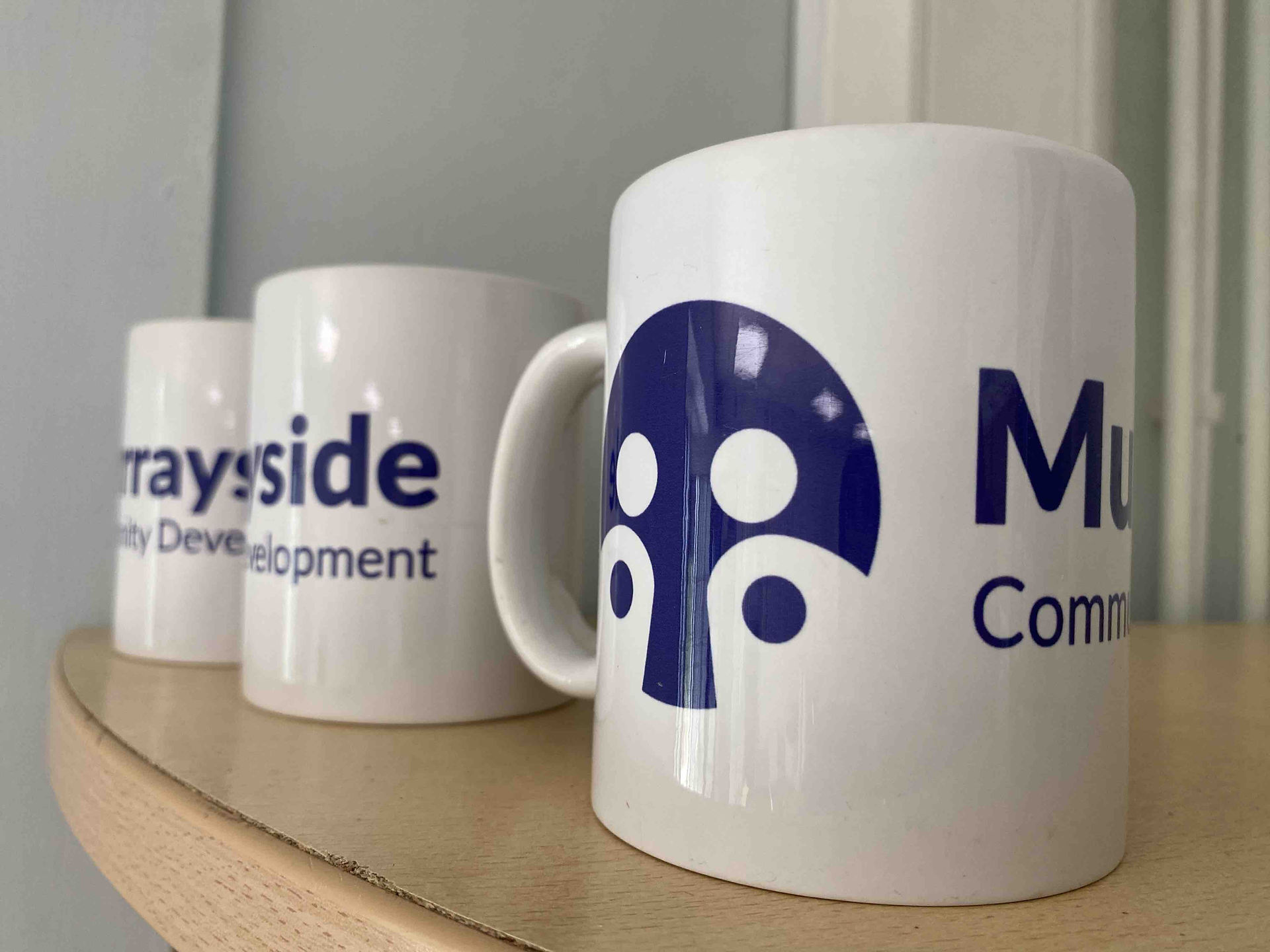 Shelf with three white mugs on it. Mugs have Murrayside Development Centre written on them, along with a logo