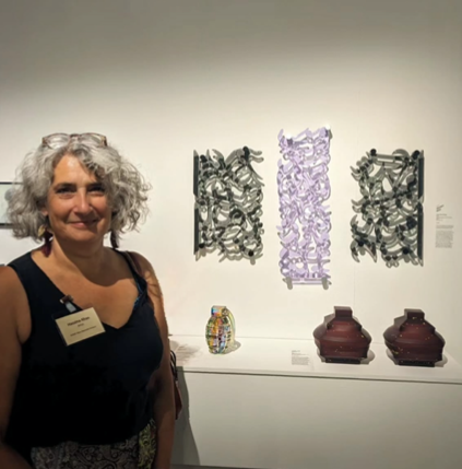 Woman in a black vest standing in front of three glass wall sculptures and various other decorative items on a shelf