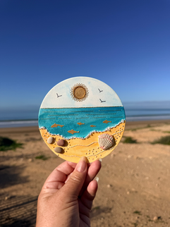 Beach scene on a sunny day with a hand holding a round piece of paper with an embroidered beach scene on it, incorporating sea shells and small stones