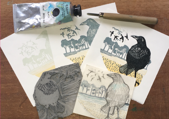 Crow-themed lino printmaking setup with carved lino block, tools, ink tube, and multiple test prints of a crow against trees and flying birds.