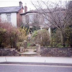 Brick property with other properties to the side.  Trees and hedges to the foreground, separating it from the road