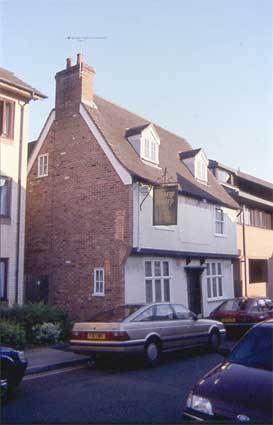 Front elevation of a house with two bottom windows, two top windows and an attic window. Between the top two windows is a blank space. Building is in a good state of repair and has a sign handing from a first floor bracket. Cars are retro