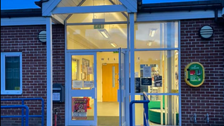 entrance to a building with a white porch and a defibrulator to the left of the door. It's dusk and the lights are on inside the building