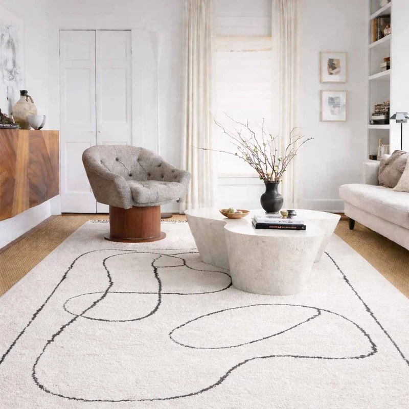 Ivory abstract line rug beneath sculptural white coffee tables, beside a grey armchair and cream sofa in a bright living room