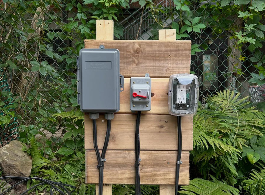 Panneau électrique avec trois boîtiers, câbles noirs et arrière-plan verdoyant LMBC ÉLECTRIQUE.