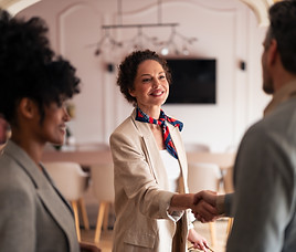 A professional woman in a beige blazer greets colleagues with a warm handshake in a bright