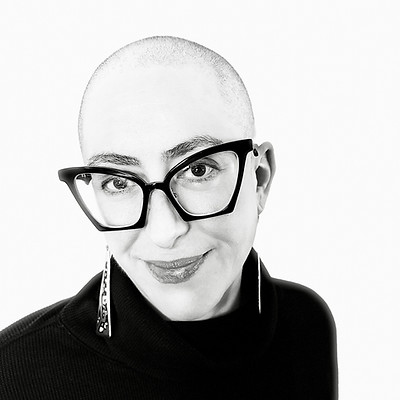 Portrait of a woman wearing dark-framed glasses and dangling earrings, against a white background.