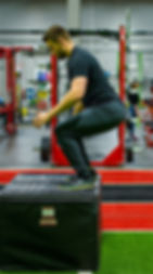 A man lands on a large black soft box after performing box jumps in a gym.