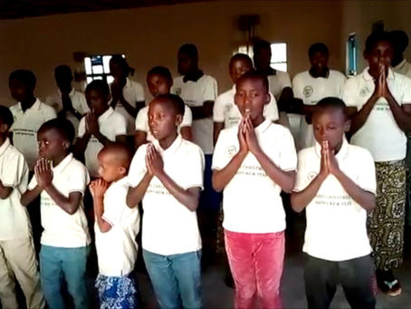 Children in white shirts stand in prayer, hands clasped, in a dim room. Mood is peaceful.
