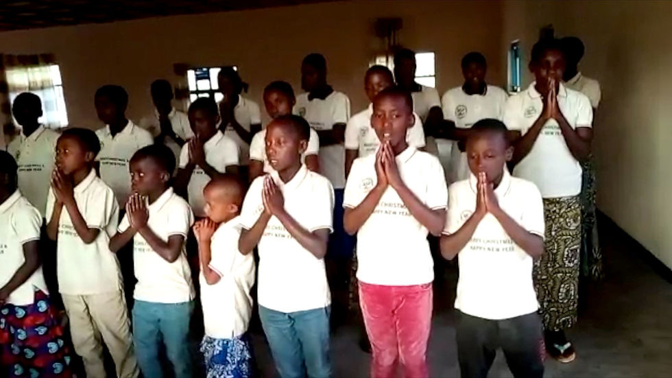 Children in white shirts stand in prayer, hands clasped, in a dim room. Some shirts display Christmas greetings. Mood is peaceful.