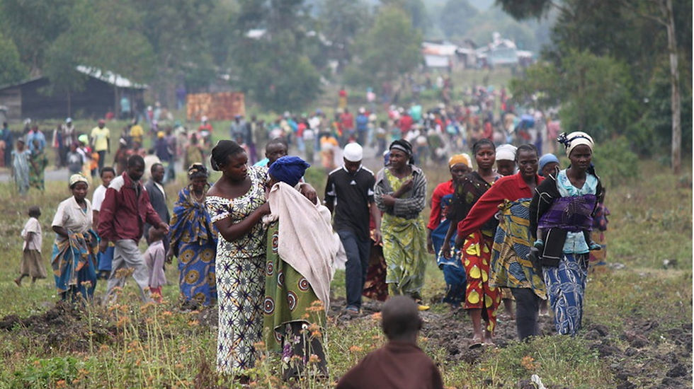 A crowd of people in colorful clothing walk through a grassy field, some carrying belongings. The mood is serious, setting is rural.