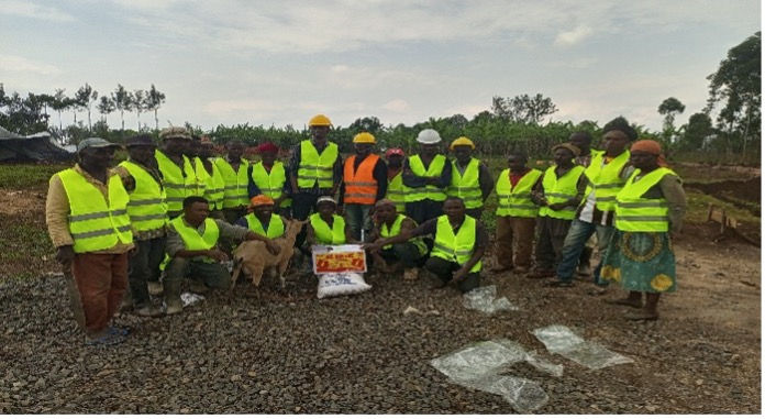 Group of people in yellow vests and helmets on a gravel surface. A goat and a bag are in front. Trees and clouds in the background.