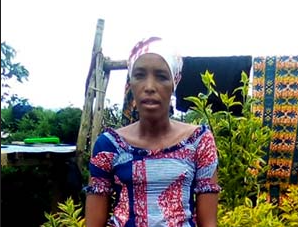 Woman, Alice, stands in a garden wearing a colorful patterned dress and headwrap. Bright yellow-green plants and patterned cloths are in the background.