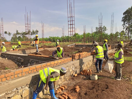 Construction workers in yellow vests build brick walls on a sunny site with metal rods and green foliage in the background.