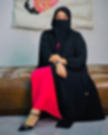 The Hijabi Realtor, Kanwal Adnan wearing a black abaya and niqab sits on a brown tufted sofa, revealing a bright pink pleated dress underneath. She has an elegant pose, with her legs crossed and hands resting on her knee, showcasing a watch and bracelet. Artwork featuring abstract shapes and floral patterns hangs on the wall behind her. A smartphone is placed on the sofa to her side. The setting appears to be an indoor, contemporary living space.