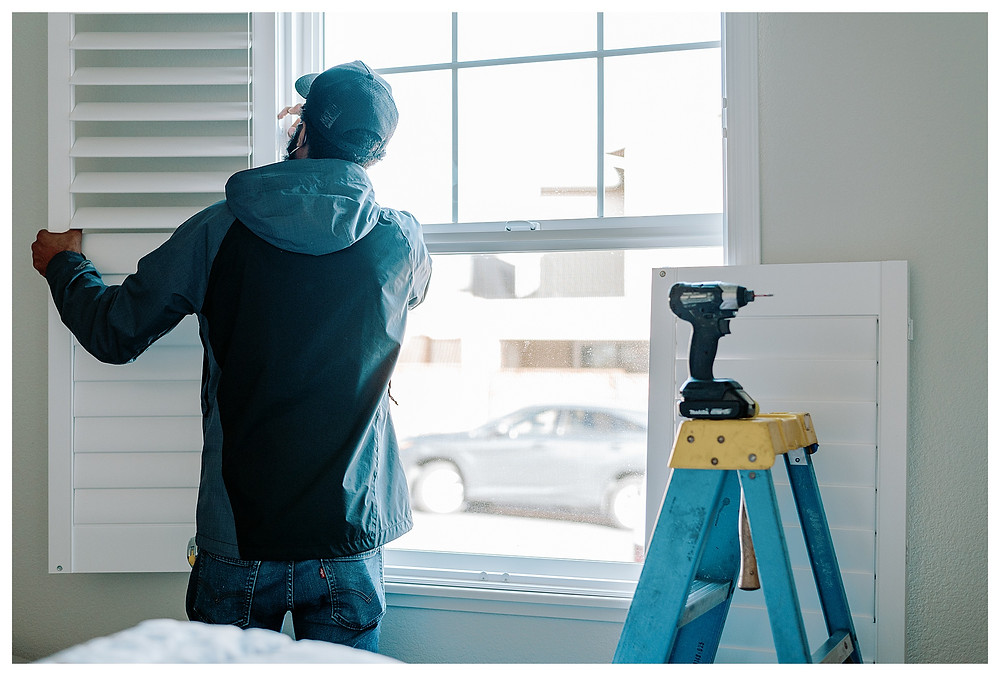A Window-ology installer hinges a shutter in place. The accompanying right shutter panel sits propped on the wall next to him, behind a blue ladder.