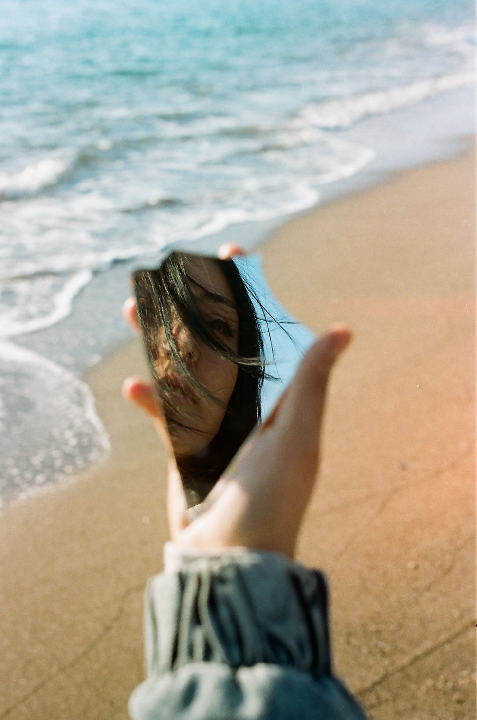 A hand holds a shard of mirror on a sandy beach, reflecting a woman's face with windswept hair. Waves gently crash in the background.