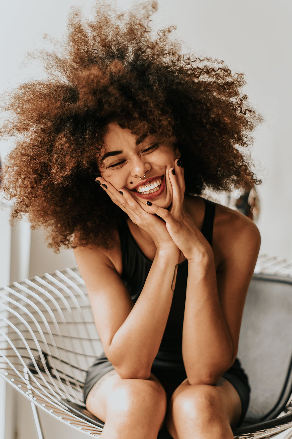 Woman with curly hair smiling, hands on cheeks, sitting on a wire chair. Light background, wearing a black top, conveying joy.