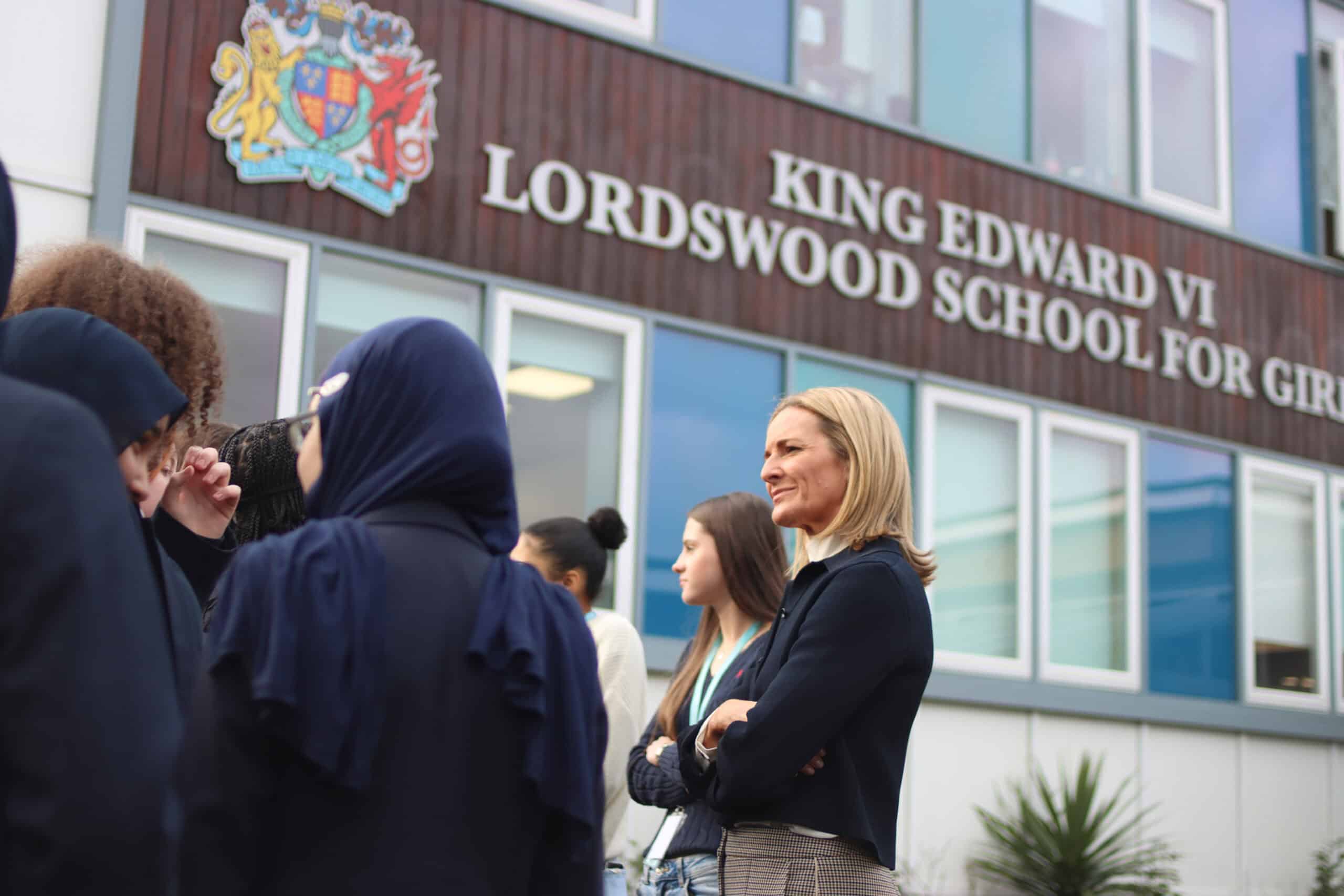 A group of female students gathers outside Lordswood School for Girls, engaged in conversation. Broadcaster and former gymnast Gabby Logan MBE stands among them, wearing a navy blazer and smiling with her arms crossed. The school's name and crest are visible on the building behind them.