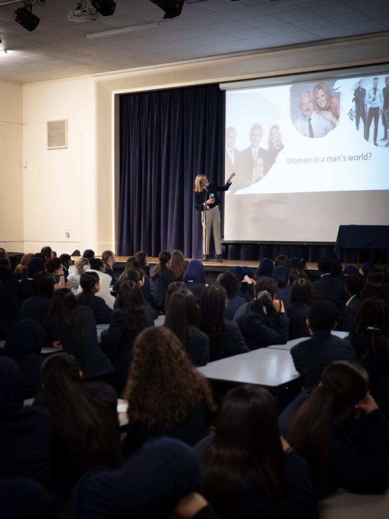 Gabby Logan MBE stands on a stage delivering an inspirational talk to a hall full of students at Lordswood School for Girls. She gestures toward a presentation slide titled “Women in a man’s world?” featuring images of male sports presenters and herself. The students, dressed in dark uniforms, sit attentively on benches and the floor, listening to her speech.
