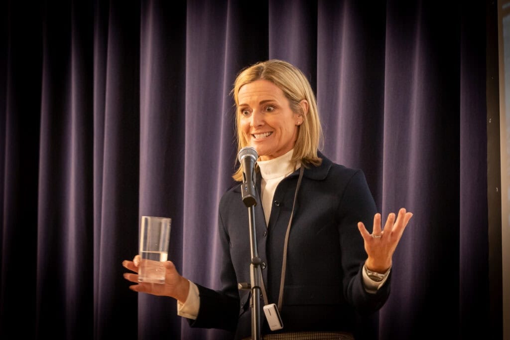 Gabby Logan MBE stands on a stage delivering an inspirational talk to a hall full of students at Lordswood School for Girls. She is holding a glass of water with her arms gesturing as she delivers an inspirational talk enthusiastically to the students.