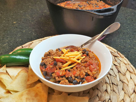 Bowl of chili with cheese, tomatoes, and beans on a woven mat. Jalapeño and tortilla chips on the side. Dark stone countertop background.