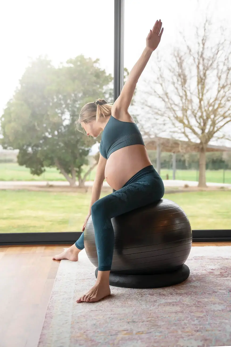 Pregnant person seated on an exercise ball reaching one arm overhead