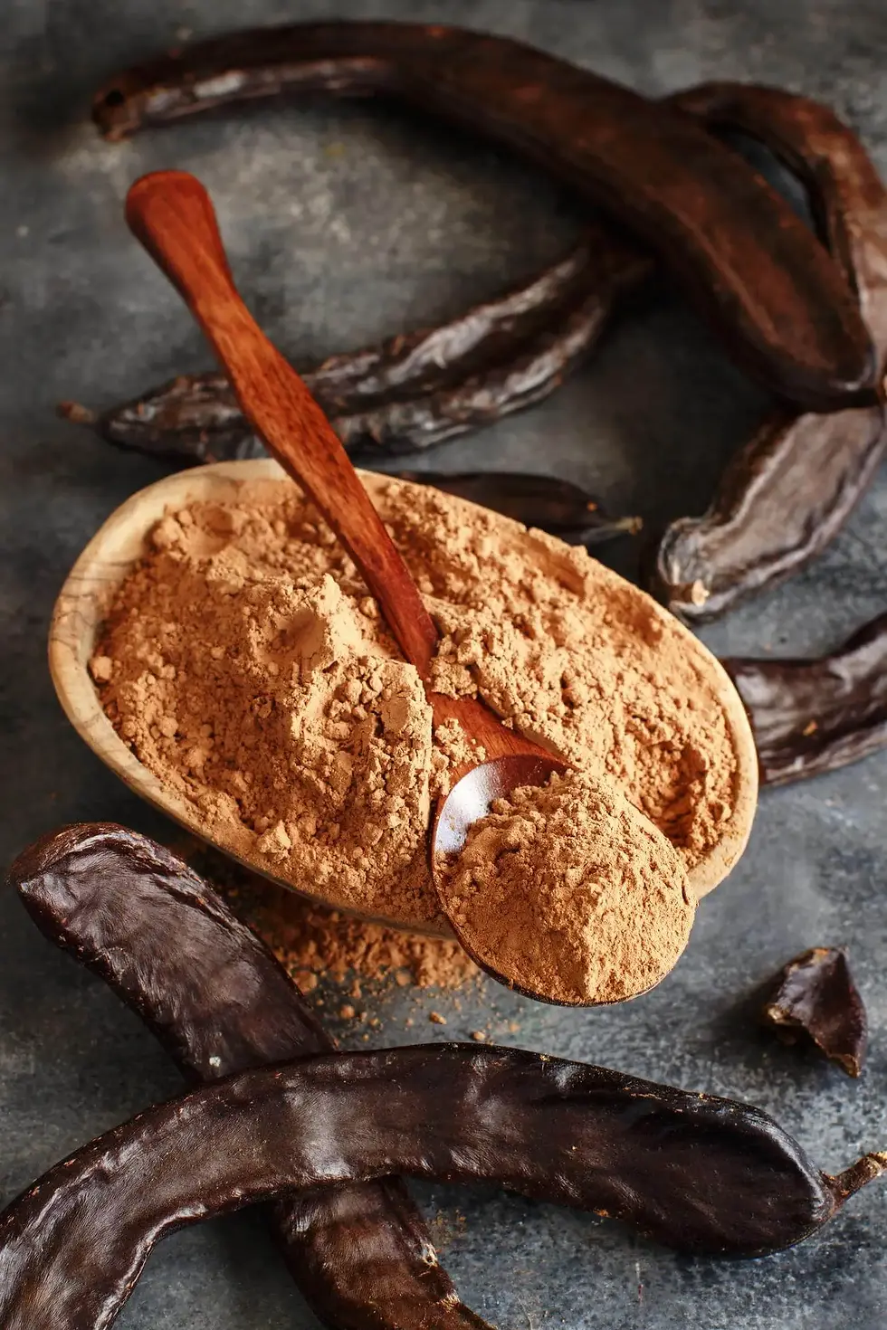 Carob powder in a bowl beside carob pods and a wooden spoon