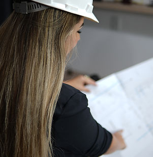 Arquiteta Jennifer Pacheco, em Itapoá, apontando para um projeto arquitetônico impresso, usando capacete de obra branco, especialista em projetos residenciais e comerciais.