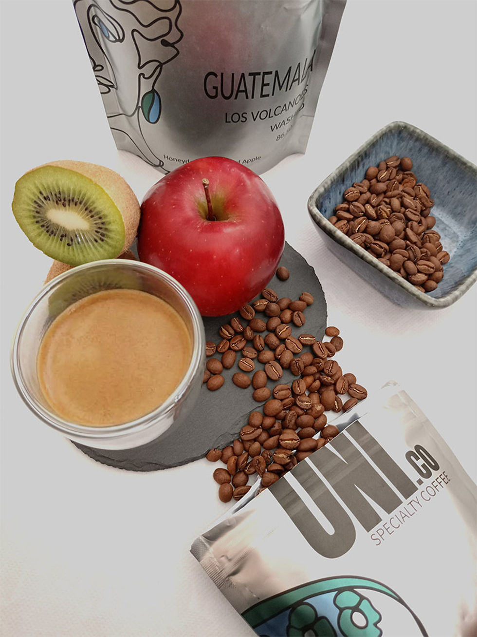 A cup of espresso next to a Uni.co coffee bag, a fresh red apple, and a sliced kiwi on a light wood table.