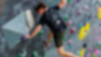 Person climbing an indoor wall with colorful holds, wearing a black shirt and green shorts. Focused and intense atmosphere.