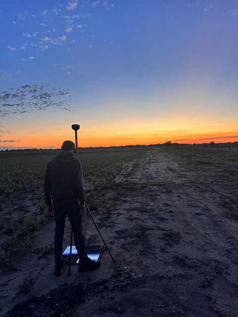 A surveyor setting up a GNSS rover pole in a farm field at sunset