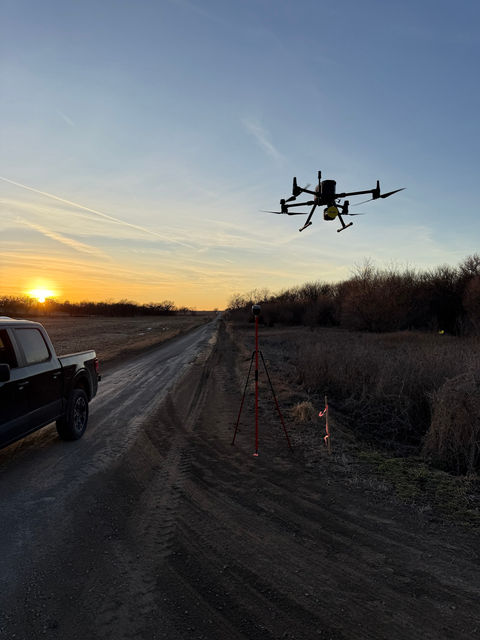 A survey drone flying at sunset above a dirt road, with a pickup truck and a GNSS tripod below