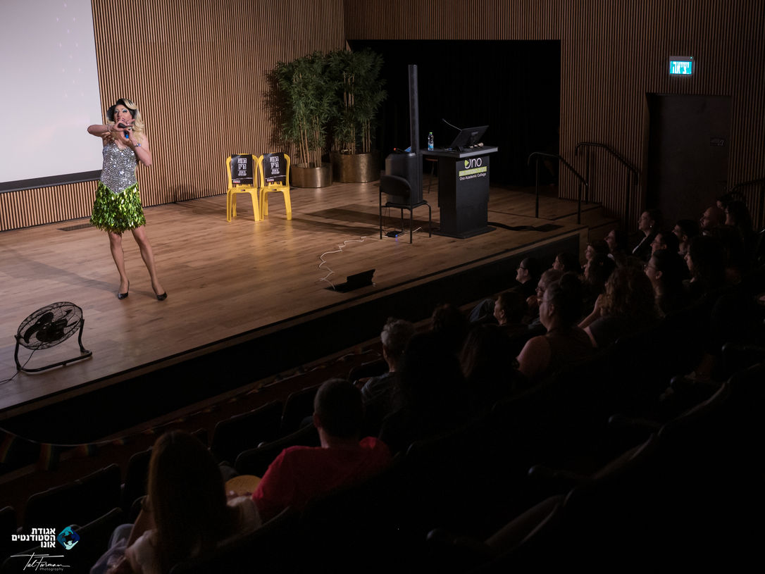 אישה לבושה בבגד יפה, מדברת למיקרופון על במה, עם קהל מאחוריה