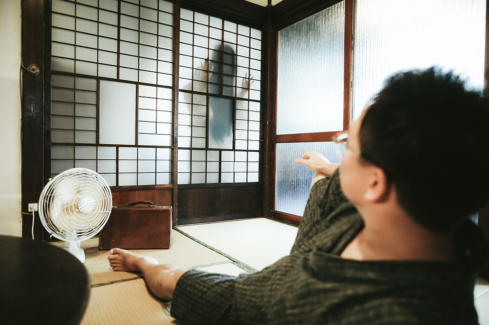 A man in a kimono relaxes in an traditional-style Japanese room, complete with an electric fan, an old suitcase, tatami mats for the floors, and sliding doors. A ghostly woman with long hair covering her face stands in front of the sliding door, her hands pressed on the paper screens.