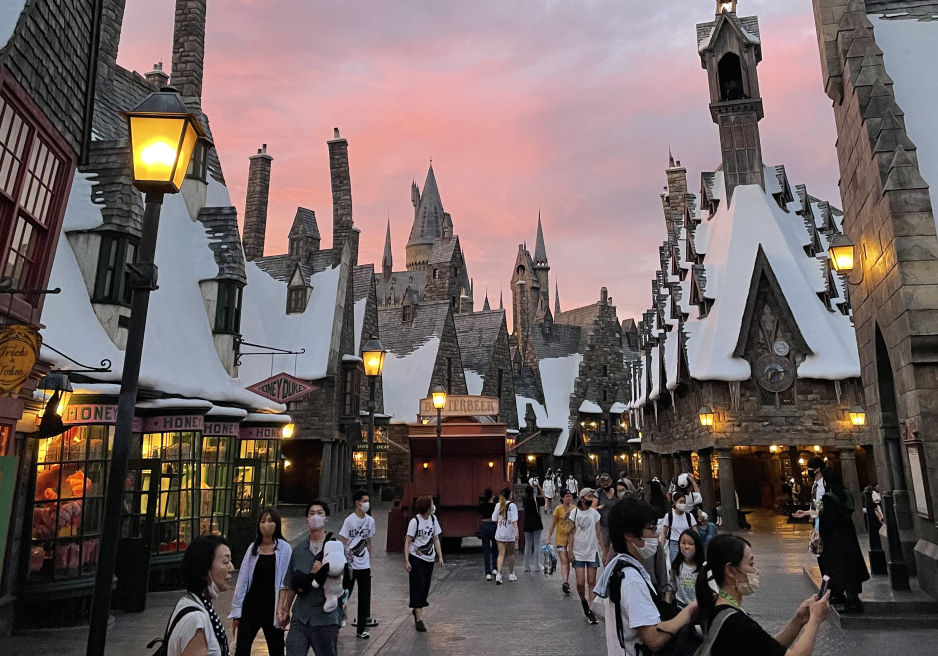 People walk through snowy Hogsmeade village at sunset, surrounded by shops with signs for Honeydukes and Butterbeer.