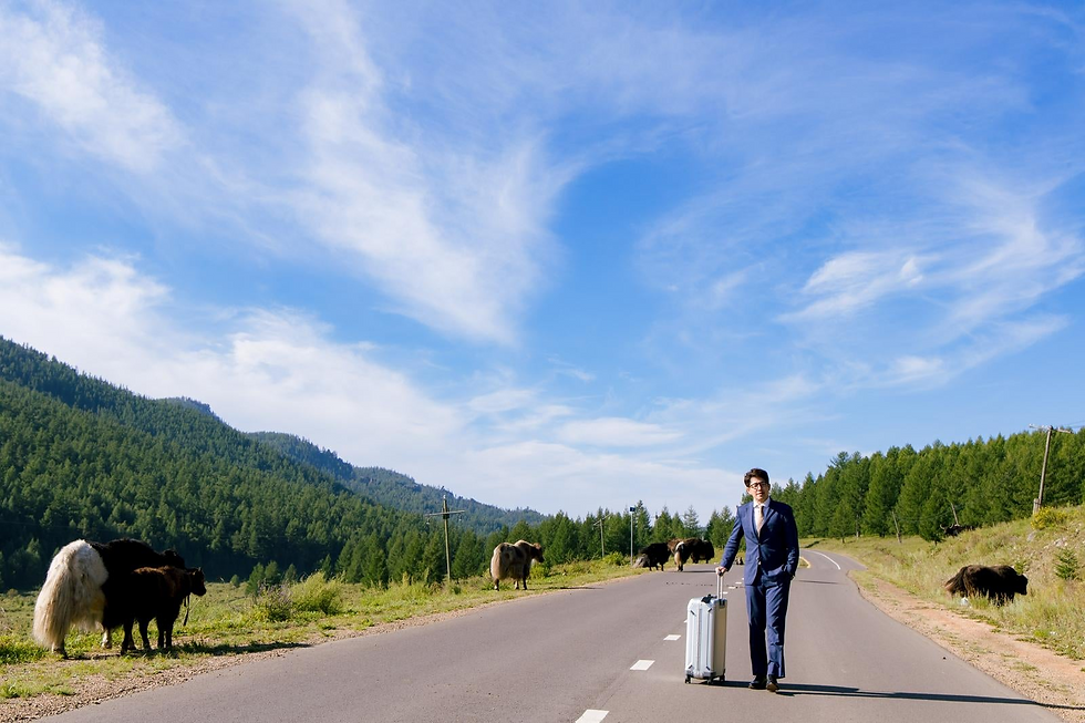 A man in a suitcase carrying a suit and looking out a place as he walks down a countryside road with mountains, greenery, and cows in the background.