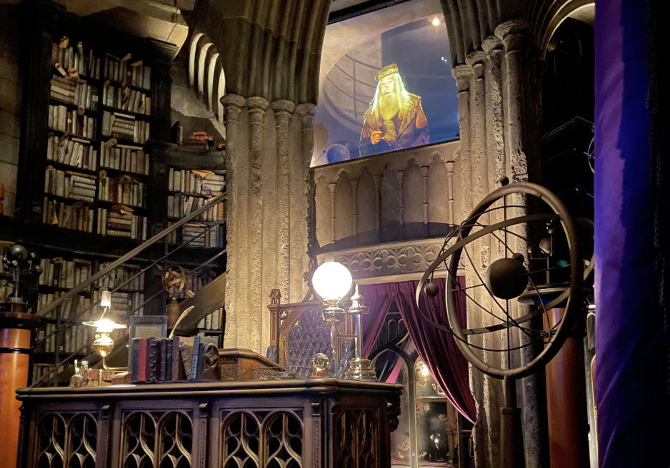 Dumbledore's office filled with bookshelves, globes, an ornate desk, purple drapes and stone arches. Dumbledore appears above on the upper level.