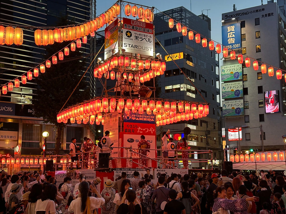 A scene from a Bon Odori festival in the early evening, featuring an elevated stage called a yagura that is surrounded by orange-red lanterns. Performers in kimonos and happi coats stand on stage as, below, spectators gather around the yagura.