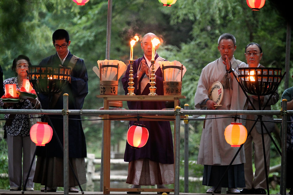 Five people, including three men in Buddhist robes, at an Obon ceremony. They stand in prayer in front of what seems to be an altar with two candles and two buckets.