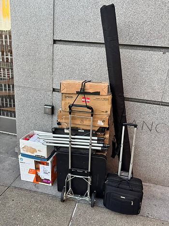 Boxes and luggage stacked on a sidewalk against a gray stone wall, including Epson and HP labeled boxes, a folded cart, and a suitcase. photo of courtroom equipment rental