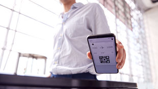 Person in a light shirt holds a phone displaying a boarding pass with FCO to LGW, in a modern, brightly-lit airport setting.