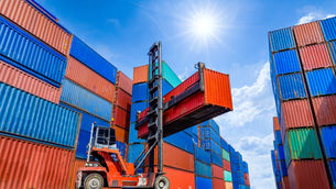 Forklift lifting red shipping container among stacked blue and orange containers under a bright sun and blue sky. Industrial setting.