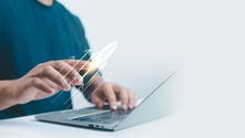 Hand touches digital rocket on screen, symbolizing growth or launch, with open laptop nearby. Blue shirt, white background.