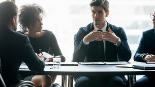 Four professionals in a meeting room discuss around a table, with notebooks and glasses of water. Large windows suggest an office setting.
