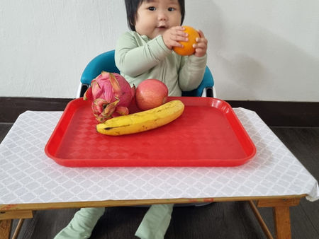 A happy child enjoys holding an orange at a table filled with fresh fruits.