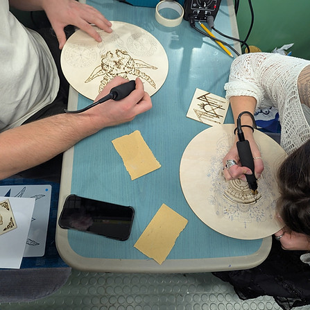Two people use pyrography tools to burn intricate designs onto wooden plates
