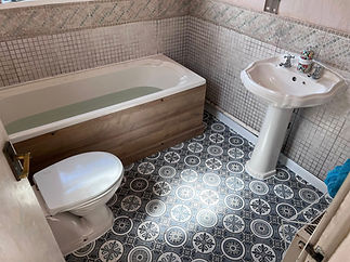 Small bathroom with patterned tile floor, white toilet, pedestal sink, and a wooden-panel bathtub partially filled with water