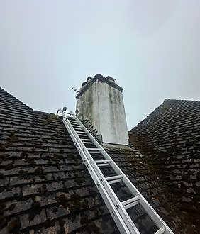 A roof ladder resting on a very mossy tiled roof leading up to a weathered chimney stack