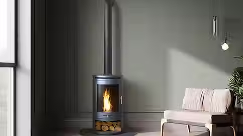 Tall charcoal grey wood-burning stove with a long flue pipe and logs stored in the base against a sage wall